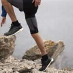 Climbers feet in black athletic shoes step along jagged rocks with a calm body of water in the background