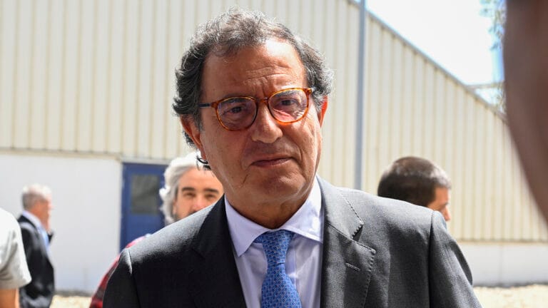 Man in a gray suit and blue tie wearing orange brown glasses outdoors facing the camera