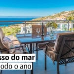 Balcony seating area with a glass table and four cushioned chairs overlooking the sea and hillside houses captioned A um passo do mar durante todo o ano
