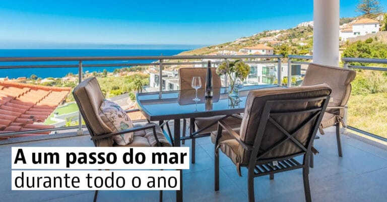 Balcony seating area with a glass table and four cushioned chairs overlooking the sea and hillside houses captioned A um passo do mar durante todo o ano