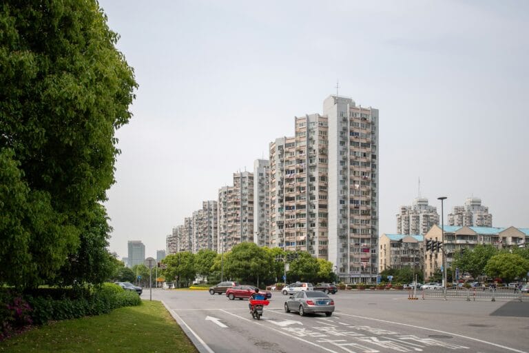 Tree lined city street with tall residential buildings on the right and several cars in the road