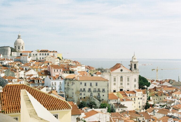 Coastal town with white buildings and red tiled roofs a large domed church on the left and the sea in the distance under a blue sky