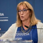 Woman with glasses speaks at a lectern during a government press conference in Portugal backdrop reads República Portuguesa