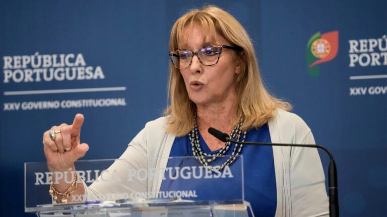 Woman with glasses speaks at a lectern during a government press conference in Portugal backdrop reads República Portuguesa