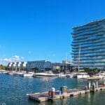 Marina scene with moored yachts and a curved glass faced residential tower on the waterfront under a clear blue sky