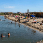 Busy riverside beach with people swimming and sunbathing on a rocky shore under a blue sky a red flag on a building in the background