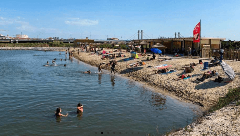 Busy riverside beach with people swimming and sunbathing on a rocky shore under a blue sky a red flag on a building in the background