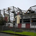 Ruined industrial building with collapsed roof and twisted metal framework surrounded by a chain link fence