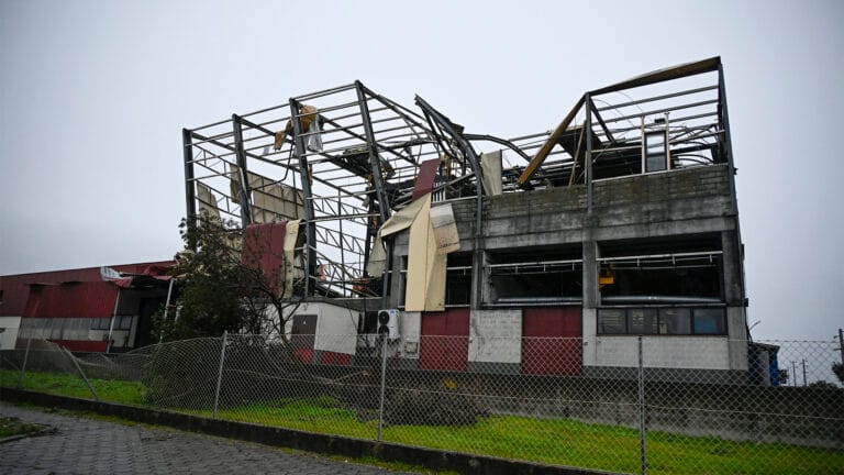 Ruined industrial building with collapsed roof and twisted metal framework surrounded by a chain link fence