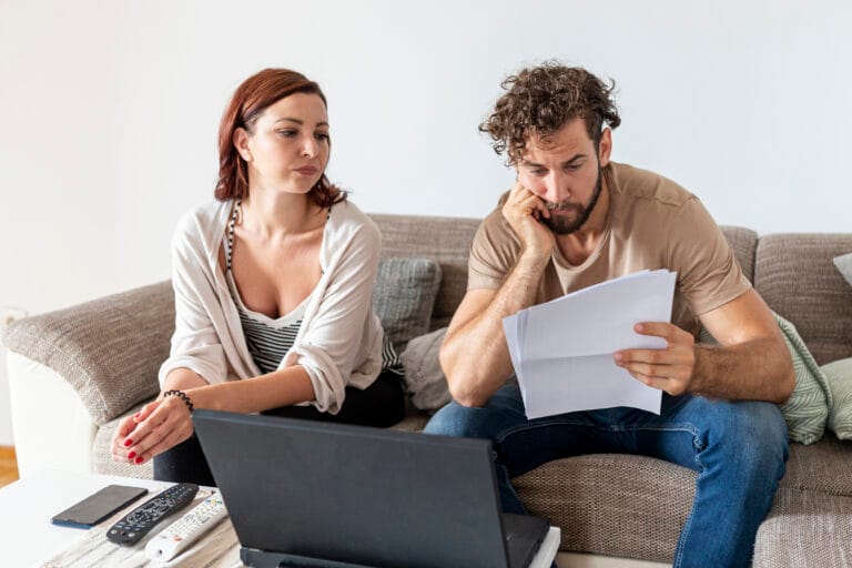 Man on a sofa studying a stack of papers while a laptop rests on the coffee table nearby with a woman seated beside him in a living room setting