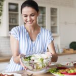 Smiling woman in a blue and white striped shirt holds a bowl of salad in a bright kitchen