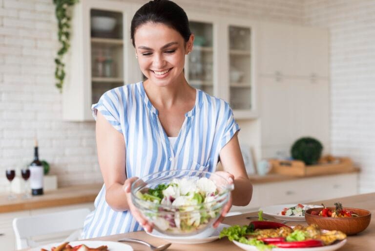 Smiling woman in a blue and white striped shirt holds a bowl of salad in a bright kitchen