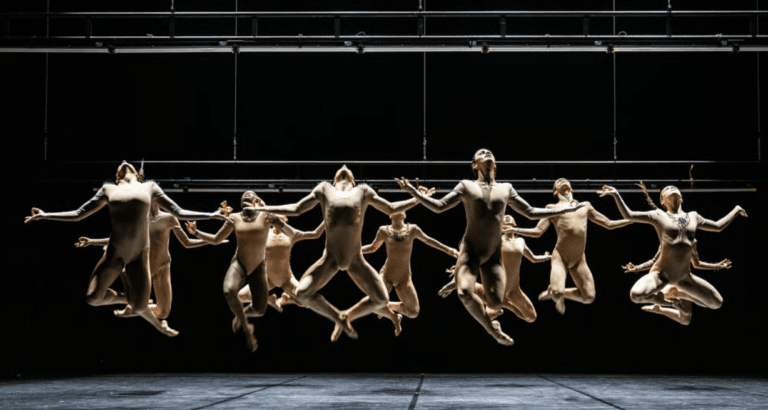 Group of dancers in beige unitards performing a synchronized jump on a dark stage mid air with arms extended