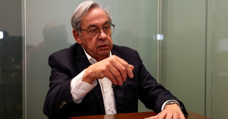 Older man in a dark blazer and white shirt sits at a wooden table gesturing with his hands during a discussion in a glass walled office