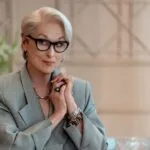 Older woman with short gray hair and black glasses in a light gray blazer hands clasped at a table
