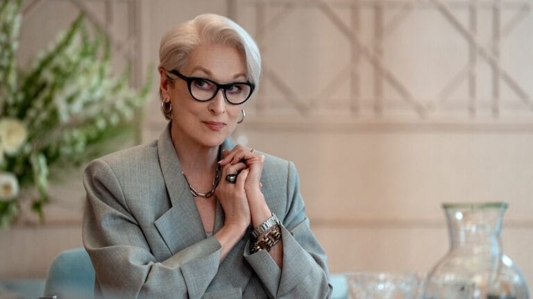 Older woman with short gray hair and black glasses in a light gray blazer hands clasped at a table