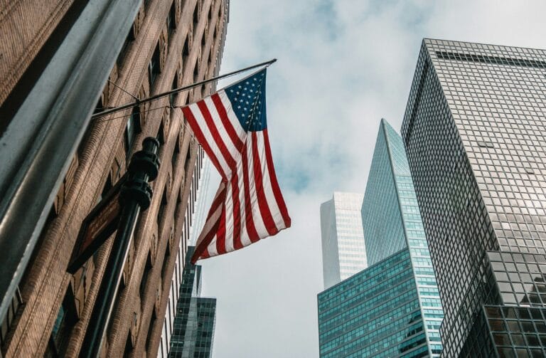 American flag waving between tall brick and glass skyscrapers in a city street under a cloudy sky