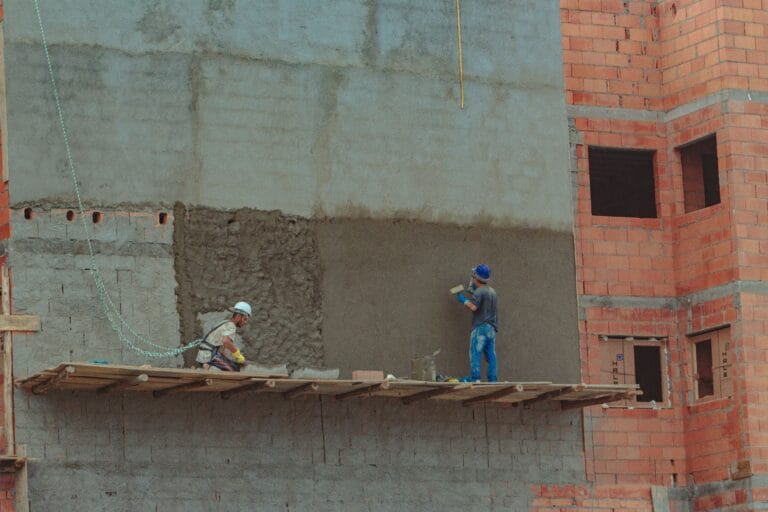 Two construction workers in hard hats stand on a wooden scaffold against a partially finished concrete wall and an orange brick building under construction
