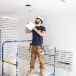 Construction worker on scaffolding inside a partially finished room wearing a hard hat and safety glasses holding a clipboard and inspecting wires in the ceiling