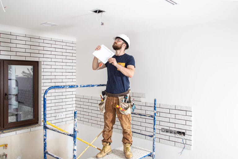 Construction worker on scaffolding inside a partially finished room wearing a hard hat and safety glasses holding a clipboard and inspecting wires in the ceiling