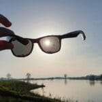 Hand grips sunglasses against the bright sun over a calm lakeside landscape