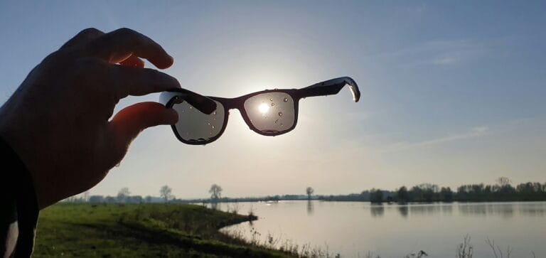 Hand grips sunglasses against the bright sun over a calm lakeside landscape