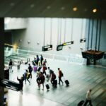 Busy airport terminal with travelers walking in a group pulling rolling suitcases along a wide tiled concourse under hanging signs