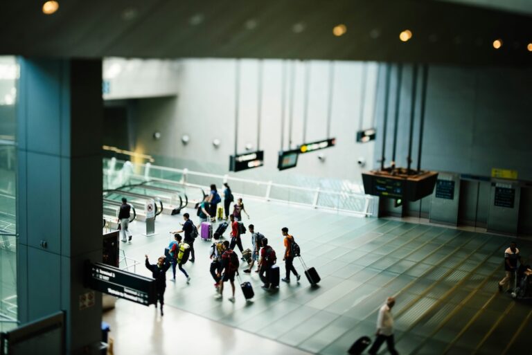 Busy airport terminal with travelers walking in a group pulling rolling suitcases along a wide tiled concourse under hanging signs