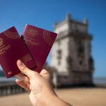 Two red Portugal passports held up in the foreground with a blurred seaside fortress in the background
