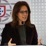 Woman with glasses speaks at a podium gesturing with her hands in front of a banner featuring a Portuguese emblem