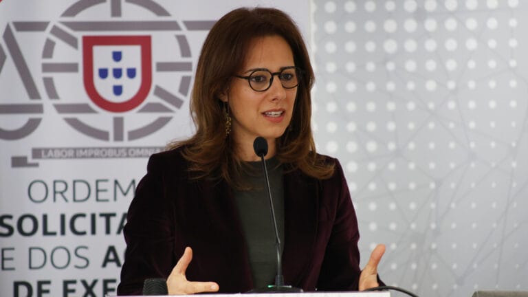 Woman with glasses speaks at a podium gesturing with her hands in front of a banner featuring a Portuguese emblem
