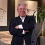 Older man in a navy suit with arms crossed in a modern office lounge with plant and architectural drawings on the wall behind him