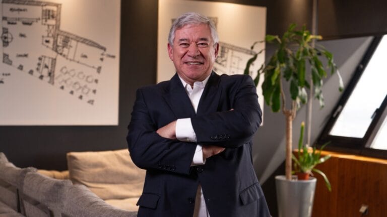 Older man in a navy suit with arms crossed in a modern office lounge with plant and architectural drawings on the wall behind him