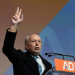 Man at a podium raises two fingers in a victory gesture while speaking into microphones with an orange AD Aliança Democrática sign in front of him