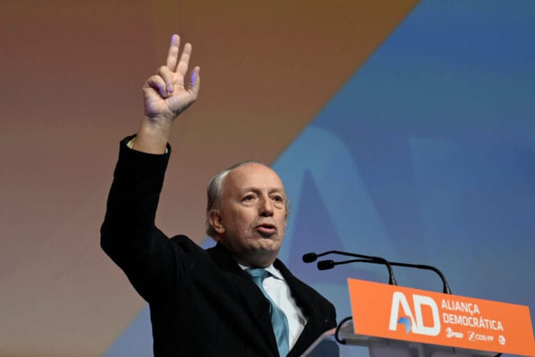 Man at a podium raises two fingers in a victory gesture while speaking into microphones with an orange AD Aliança Democrática sign in front of him