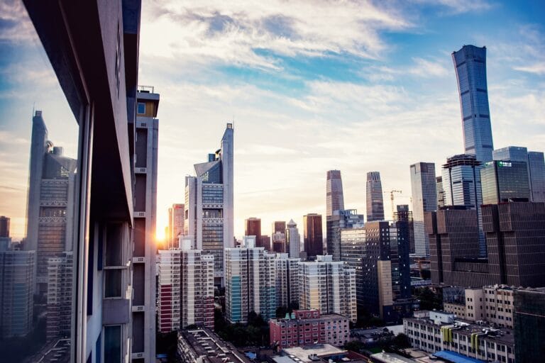 Sunset over a dense city skyline with tall glass skyscrapers and a foreground building reflecting the warm light of the setting sun on the left