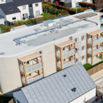 Aerial view of a curved modern apartment building with wooden framed balconies and a flat rooftop with pipes and vents