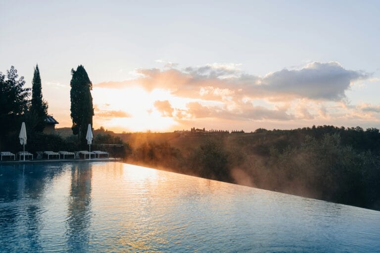 Sunset over a poolside with lounge chairs and umbrellas tall trees on the left and rolling hills in the distance