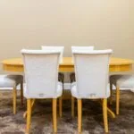 Six beige upholstered dining chairs surround a round honey colored wooden table in a neutral room with a patterned rug