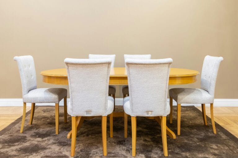 Six beige upholstered dining chairs surround a round honey colored wooden table in a neutral room with a patterned rug