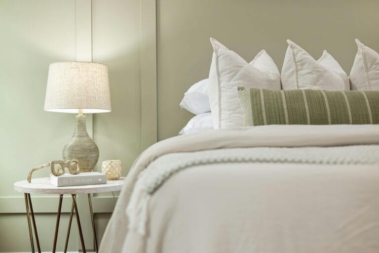 Bedside table scene woven lamp round marble table and decorative items beside a neatly made bed with white pillows and a green striped throw rack