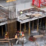 Construction site with workers in orange vests and hard hats assembling steel reinforcement framework for a building
