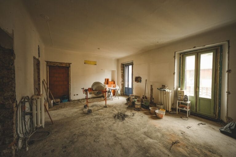 Interior of a room under renovation with a small cement mixer on wheels buckets and scattered tools on a dusty floor near a green door and window