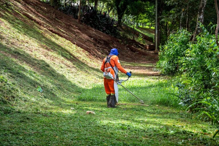 Worker in orange protective suit and blue hood using a grass trimmer to clear a grassy slope in a park lined area