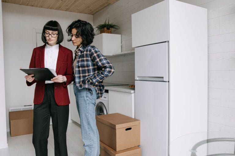Two people stand in a bright kitchen one in a red blazer showing a clipboard to the other surrounded by moving boxes on the floor and a white refrigerator nearby