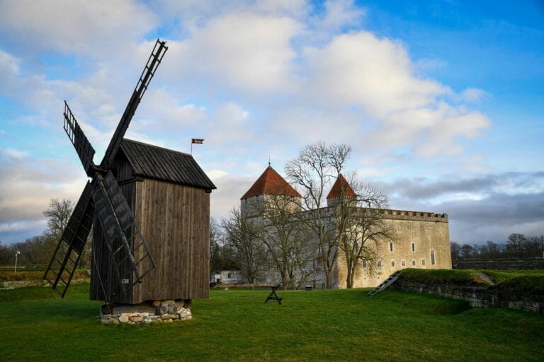 Wooden windmill in the foreground with a stone castle and leafless trees under a partly cloudy sky