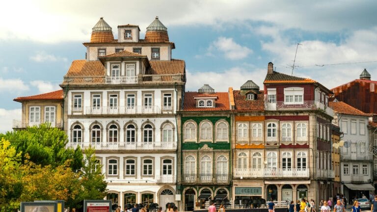 Colorful row of historic European townhouses with red tiled roofs and ornate balconies along a busy street pedestrians at ground level