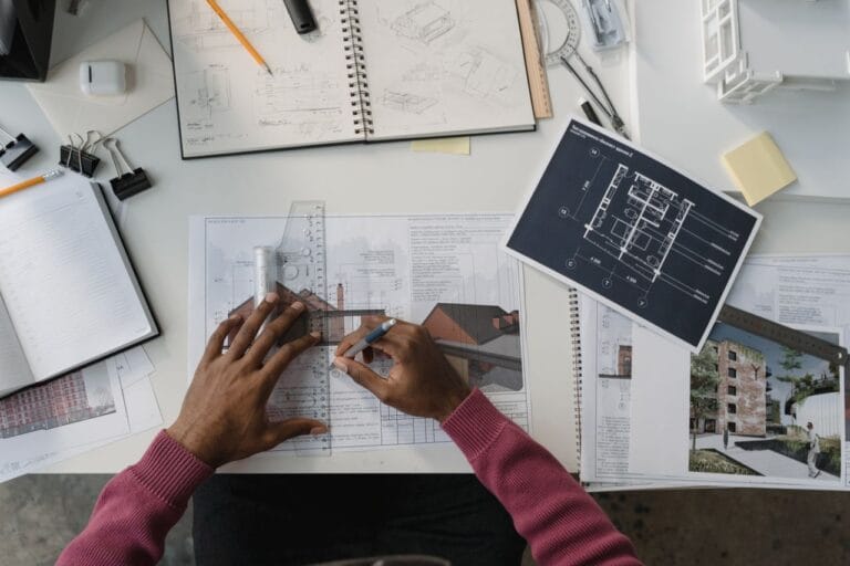 Overhead view of a designers hands measuring a blueprint with a clear ruler on a desk full of plans and sketches in progress