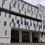Front view of a modern white building with many narrow windows a Polícia Judiciária sign is near the entrance with flagpoles to the right and a few people outside in front