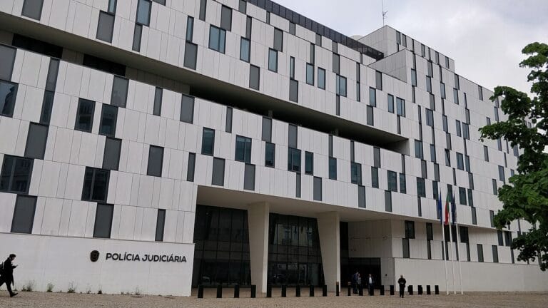 Front view of a modern white building with many narrow windows a Polícia Judiciária sign is near the entrance with flagpoles to the right and a few people outside in front